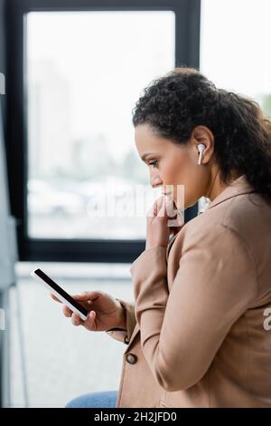 Seitenansicht einer nachdenklichen afroamerikanischen Geschäftsfrau im drahtlosen Kopfhörer, die während eines Videoanrufs auf dem Smartphone denkt Stockfoto