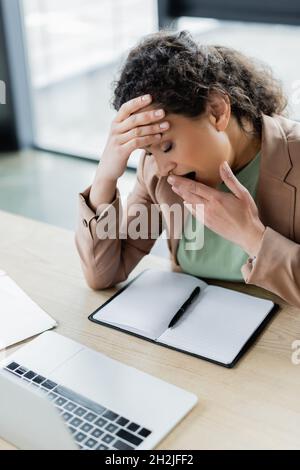 Müde afroamerikanische Geschäftsfrau gähnend mit geschlossenen Augen in der Nähe eines leeren Notizbuchs am Arbeitsplatz Stockfoto