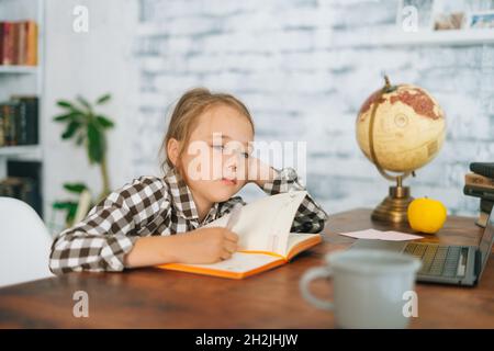 Vorderansicht der gelangweilten müde niedlichen Grundschule Kind Schulmädchen tun Schreiben Hausaufgaben halten Stift sitzen am Tisch mit Laptop und Papier-Notebook zu Hause Stockfoto