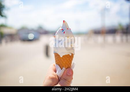 Eine weibliche Hand hält einen Eiskegel auf einem Straßenboden. Das Eis schmolz und lief mir die Finger und die Hand herunter. An einem heißen Tag draußen. Nahaufnahme, nein Stockfoto