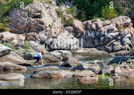 Große Steinbrocken umgeben die Ufer eines Waldflusses mit einem kalten, schnellen Wasserfluss, eine Dame sitzt am Ufer und wäscht ihre Hände im Wasser. Stockfoto