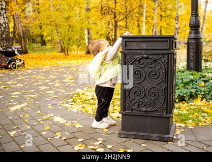 Kleine Kleinkinder wirft Müll in den Abfalleimer im Herbstpark. Einbringen kultureller Normen von Geburt an Stockfoto