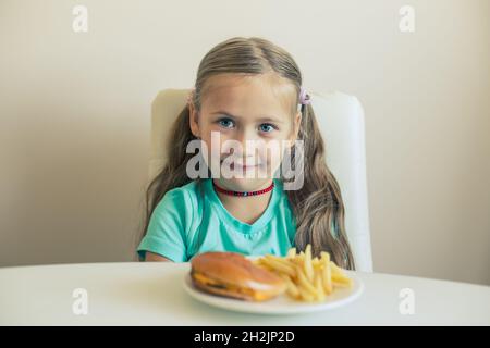 Ein lächelndes kleines Mädchen sitzt am Tisch mit Hamburgern und Pommes Frites Stockfoto