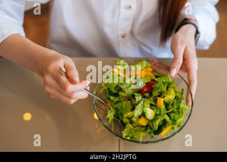 Draufsicht auf weibliche Hände, die Salat mit grünen Gurken, roten Tomaten und Salat in einer Schüssel mit Gabel am Holztisch in der modernen Küche mischen. Stockfoto