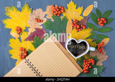 Herzförmige Tasse schwarzen heißen Kaffees mit Dampf, leeres liniertes Notizbuch und Herbstblättern auf blauem Holzhintergrund. Herbstdekor, Herbststimmung, Herbststimmung Stockfoto