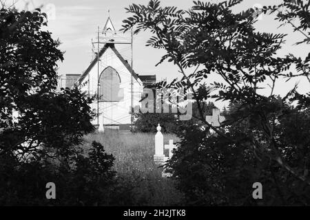 Eine alte Kirche, die repariert wird, mit Gerüsten, die um sie herum auf einem alten, überwucherten Friedhof errichtet wurden. Stockfoto