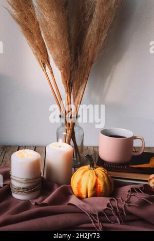 Gelber Kürbis, Kaffeetasse, Kerzen und Pampagras in Glasvase. Stillleben im Herbst. Einrichtung im Boho-Stil. Stockfoto