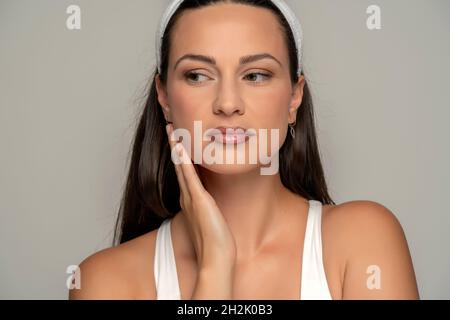 Eine junge Frau mit einem leichten Make-up berührt ihr Gesicht auf einem grauen Hintergrund Stockfoto