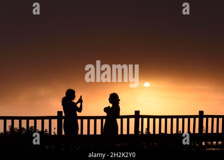 Silhoutte von zwei jungen erwachsenen Frauen, die bei Sonnenuntergang Sonne stehen. Sie machen Fotos Stockfoto