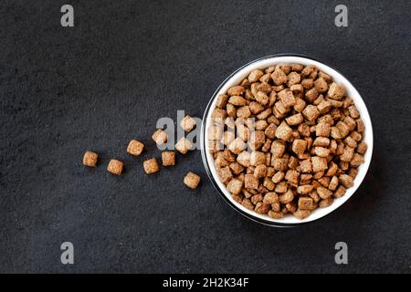 Trockenes Tierfutter in einer weißen Keramikschale auf schwarzem Hintergrund. Flach liegend, Draufsicht, Kopierbereich Stockfoto