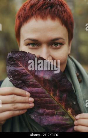 Eine Frau mittleren Alters von 30-40 Jahren mit einem herbstlichen burgunderroten Blatt, ihr Gesicht ist bedeckt. Nahaufnahme Porträt, junge Erwachsene Frau Stockfoto