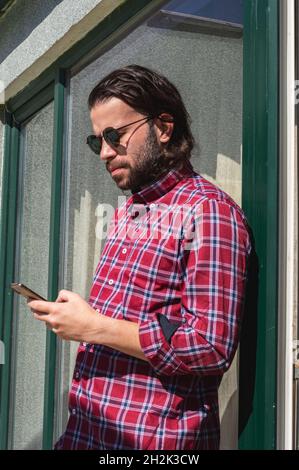 Porträt eines bärtigen jungen kaukasischen erwachsenen Mannes, der mit einem roten karierten Hemd eine Nachricht auf das Mobiltelefon eintippt Stockfoto