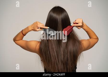 Rückansicht einer Frau, die ihr Haar mit zwei Haarbürsten auf grauem Hintergrund putzt Stockfoto