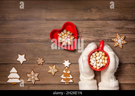 Gemütliche Tasse heißer Schokolade in einem roten Becher, gehalten von cremefarbenen Fäustlingen auf einem rustikalen Holztisch mit weihnachtlichen Schneeflocken-Keksen im Winter. Flach mit Polizist legen Stockfoto