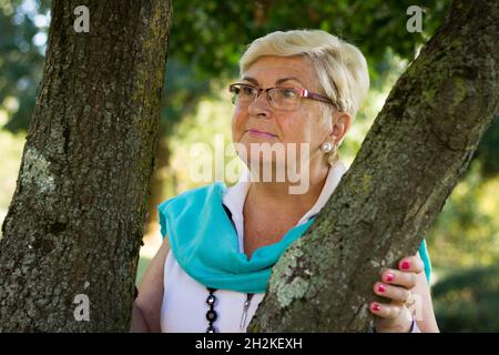 Blonde ältere Frau mit Brille posiert zwischen Baumstämmen im Park. Ältere Dame Porträt lächelt in der Natur. Rentner mit grünem Schal Stockfoto