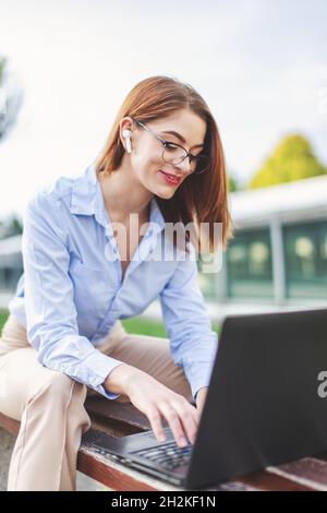 Junge Rothaarige, die online per Laptop und Freisprecheinrichtung im Park kommuniziert Stockfoto