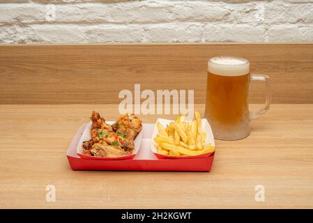 Gebratene Hähnchenflügel mit Thai-Sauce, Pommes Frites-Garnitur und ein Becher appetitlich kaltes Bier auf einem Holztisch Stockfoto