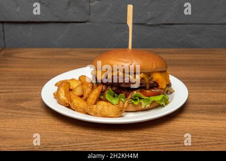 Rindfleisch-Burger vom Holzkohlegrill mit tropfendem geschmolzenem Cheddar-Käse, karamellisierten Zwiebeln, Tomaten und Senf mit Keil-Pommes Stockfoto