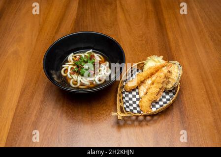 Schwarze Schüssel mit udon-Nudeln mit Gemüse und Tempura Teller mit Garnelen und Gemüse auf Korbkorb auf Holztisch Stockfoto