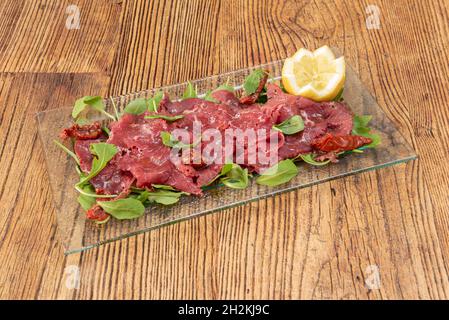 Italienisches Gericht aus rohem Rindfleisch, mariniert mit Salz und Olivenöl, bedeckt mit Rucola-Blättern und dehydrierten Tomaten mit Zitrone auf einer transparenten Glasschale Stockfoto