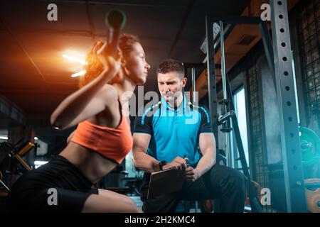 Junge Frauen trainieren und trainieren Langhantel hocken im Fitnessstudio mit Personal Trainer Stockfoto