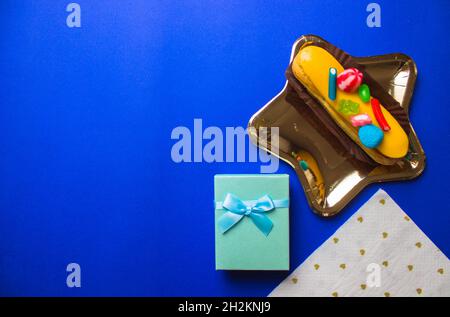 Ein goldener sternförmiger Teller mit köstlicher eclair, blauer Geschenkbox, weiße herzförmige Serviette liegen flach auf dem hellblauen Tisch. Festliche Dekoration zum Geburtstag, Stockfoto