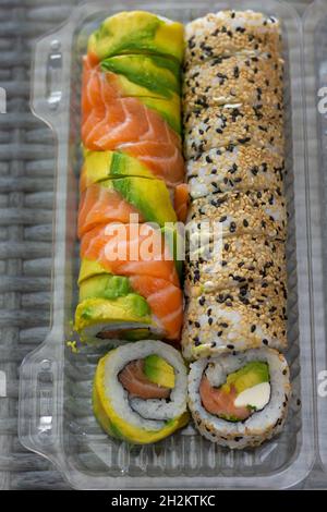 Sushi-Rollen mischen sich in Lachs, Avocado und Sesamsamen auf einem transaparenten Plastikbehälter. Food to go, Auftragslieferung, Take-away-Konzepte Stockfoto