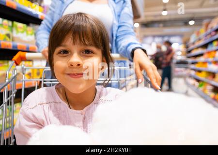 Ziemlich nettes Mädchen, das in die Kamera schaut, während sie im Einkaufswagen sitzt und ihre junge Mutter den Wagen drückt, der im Laden ist und Produkte kauft Stockfoto