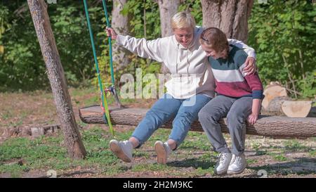 Ein Teenager-Mädchen und ihre Mutter reiten auf einer langen Holzschaukel in der Natur. Stockfoto