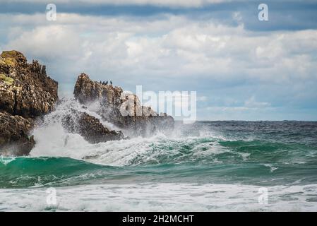 Am Felsenstrand mit wunderschönem Himmel und Wolken planschen Ozeanwasser. Im Winter plätschert die Meereswelle auf Stein an der Küste. Meereswellen schlagen gegen Felsen Stockfoto