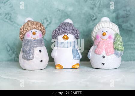 Drei Keramik-Neujahrsspielzeuge - ein Pinguin und Schneemänner in gestrickten Hüten und Tüchern auf türkisfarbenem Hintergrund. Neujahr und weihnachtskonzept. Begrüßung Stockfoto