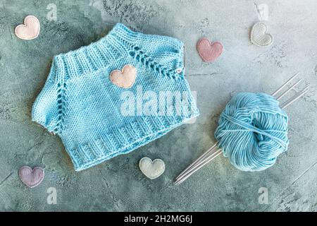 Das Konzept des Strickens von Kleidung für Spielzeug und Babys. Ein gestricktes T-Shirt aus türkisfarbenem Baumwollgarn. Eine Kugel aus Garn, Stricknadeln und Samt Stockfoto