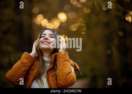 Frau lächelt mit geschlossenen Augen und Gesicht zum Himmel mit Kopfhörern und orangefarbenem Mantel im Park bei Sonnenuntergang Stockfoto