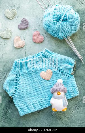 Das Konzept des Strickens von Kleidung für Spielzeug und Babys. Ein gestricktes T-Shirt aus türkisfarbenem Baumwollgarn. Eine Kugel aus Garn, Stricknadeln, Spielzeug und Samt Stockfoto