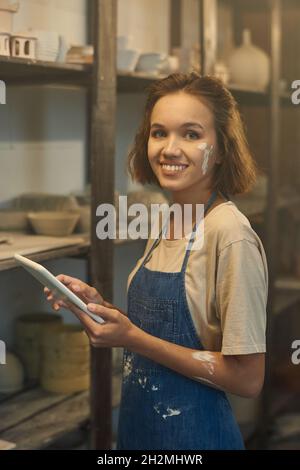 Porträt einer glücklichen jungen Töpferin in Denim-Schürze mit digitaler Tablette im Töpferwarenlager Stockfoto