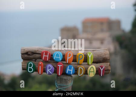 Happy Birthday Text komponiert mit mehrfarbigen Steinbuchstaben auf Holz mit altem Dorf auf der Rückseite des Meeres Stockfoto