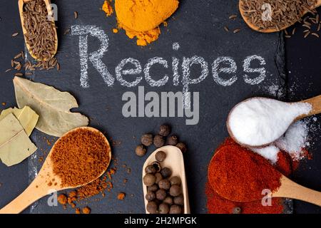 Rezepte, die mit weißer Kreide auf einer schwarzen Steinfliese geschrieben wurden, umgeben von verschiedenen Gewürzen auf Holzlöffeln. Curcuma, Curri, Paprika, Lorbeerblätter, Meersalz Stockfoto