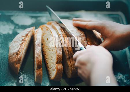 Ein Mann mit einem scharfen Messer schneidet frisches, rundes Weizenbrot, das auf einem Backblech liegt, in Scheiben. Hausmannskost. Gebäck zum Frühstück. Stockfoto