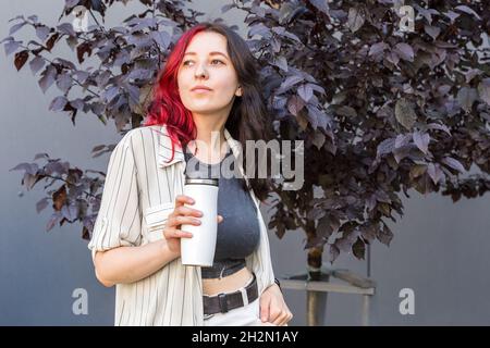 Junge Frau mit kreativen schwarzen und roten Haaren hält einen weißen, wiederverwendbaren, isolierten Becher, damit ihr Kaffee in die Hände kommt. Stockfoto
