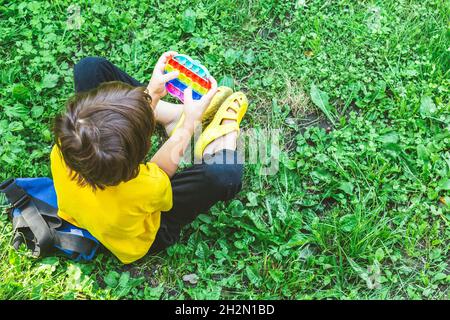 Niedlicher Vorschulkinder spielt mit dem Pop IT Flexible Sensory Fidget-Spielzeug, das draußen auf dem Gras sitzt. Aktivitäten für Kinder, geistige Entwicklung. Selektiv Stockfoto