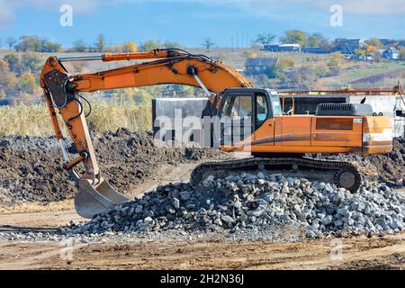 Ein Bau-Raupenbagger klettert während des Baus einer Straße auf einer ländlichen Straße am Mittag auf einen Haufen Pflastersteine. Stockfoto