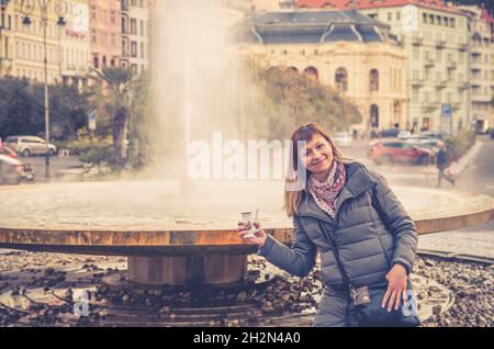 Junge Frau Tourist mit grauer Jacke posiert und halten therapeutische Mineralwasser in der Tasse in der Nähe Brunnen heißen Quelle Geysir Vridlo in Karlsbad historica Stockfoto