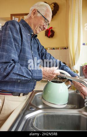 Lächelnder Älterer Mann, Der Am Waschbecken Stand Und Den Wasserkocher Füllte, Um Einen Heißen Drink Zu Machen Stockfoto