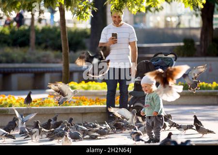 Lächelnder Vater fotografiert das Kind, das im Park unter Tauben spielt Stockfoto