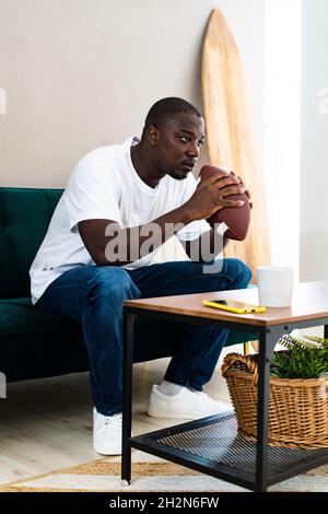 Mann hält Rugby Ball, während er zu Hause auf dem Sofa sitzt Stockfoto