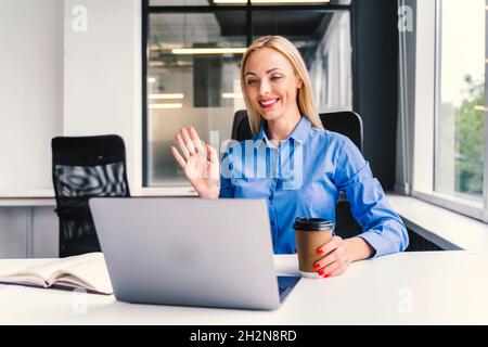 Lächelnde blonde Geschäftsfrau winkt während eines Videoanrufs über den Laptop im Büro mit der Hand Stockfoto