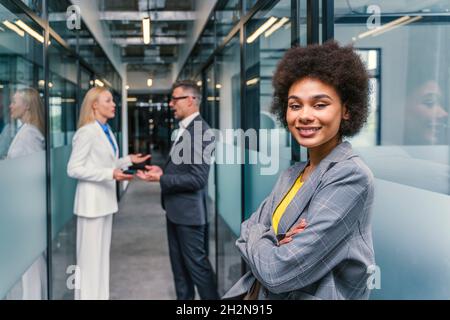 Junge Geschäftsfrau, die mit gekreuzten Armen steht, während eine Kollegin und eine Kollegin im Korridor diskutieren Stockfoto