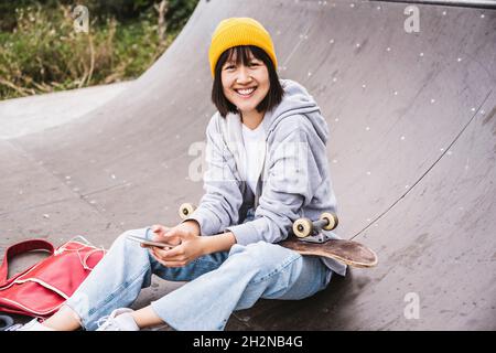 Lächelndes Teenager-Mädchen mit Strickmütze, das Smartphone hält, während es im Skateboard-Park sitzt Stockfoto