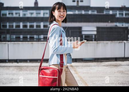 Lächelndes Mädchen im Teenageralter, das eine Schultertasche mit einem Smartphone trägt, während es auf dem Dach geht Stockfoto