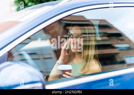Lächelnde Frau mit Kaffeetasse, die im Auto auf dem Smartphone spricht Stockfoto